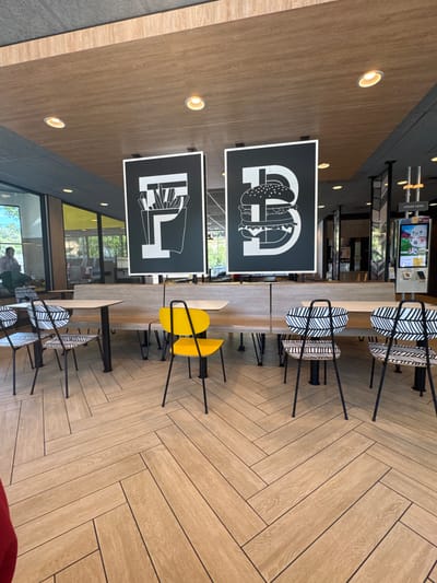 This is an eye-level, medium shot of the interior of a McDonald's restaurant, likely located in Placerville, United States of America, as suggested by the context. The scene features several dining tables and chairs set against a backdrop of a modern, clean interior.

Dominating the upper portion of the image are two large, black framed signs with white graphic representations. The left sign depicts a stylized box of french fries, and the right sign shows a large hamburger. These graphics are centrally positioned and draw the viewer's eye.

In the foreground and mid-ground, there are multiple dining tables and chairs. One table is a long, light-colored bench style table with dark metal legs. In front of this bench are several chairs. To the left, there are two chairs with black and white patterned upholstery, featuring a geometric leaf-like design. Adjacent to these, a single bright yellow chair with a distinctive backrest stands out. Further to the right, more chairs with the same black and white patterned upholstery are visible, positioned in front of another section of the bench table.

The floor is covered in light brown, wood-look tiles laid in a herringbone pattern, creating a visually dynamic texture. The ceiling is made of light wood panels, with recessed circular lights providing illumination.

In the background, a counter area is partially visible, with what appear to be menu boards or digital displays. To the far right, a self-order kiosk is present, showing colorful graphics of food and drinks, including a cartoon character. Natural light streams in from windows to the left, revealing a glimpse of greenery and people outside, suggesting a daytime setting. One person, wearing a light pink shirt, is seated at a table on the far left, facing away from the camera. The overall atmosphere is clean, contemporary, and typical of a fast-food restaurant dining area.