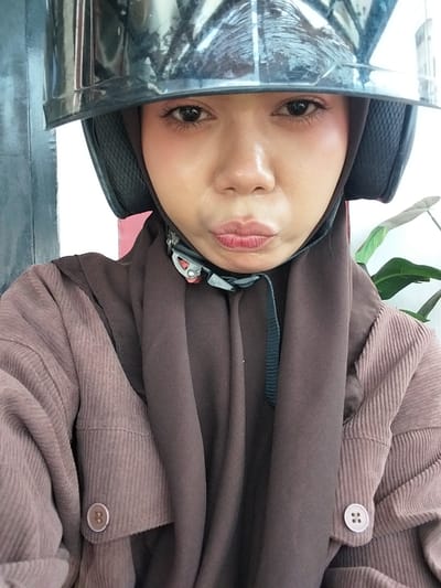 A young woman wearing a brown hijab and a brown corduroy jacket is pictured from the chest up, looking directly at the camera. She is also wearing a black helmet. Her lips are pursed. The image appears to be taken outdoors, with a blurry green plant visible on the right side and a gray, textured wall on the left. The woman's eyes are made up with dark eyeliner. There is a strap from the helmet visible on her chest. The background is out of focus, suggesting a shallow depth of field.