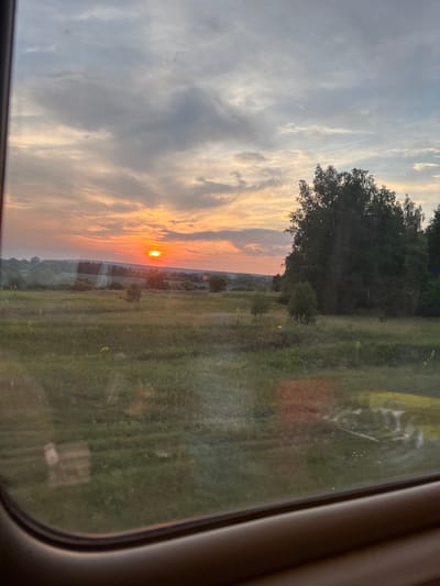 The image captures a sunset over a rural landscape, viewed through a window. The sky is a canvas of soft orange, pink, and gray clouds. The sun, a bright orange orb, is low on the horizon, casting a warm glow. Below the sky, rolling hills are visible, covered in green fields and scattered trees. A dense cluster of trees stands on the right side of the frame, their dark silhouettes contrasting with the luminous sky. The foreground shows a grassy field with a few scattered yellow flowers. The lower part of the image is framed by the dark interior of a vehicle, likely a car or bus, with a brown trim along the bottom edge. Reflections are visible on the window surface, obscuring some of the details of the landscape. The image does not contain any people or specific identifiable landmarks within Chufarovo, Russia.