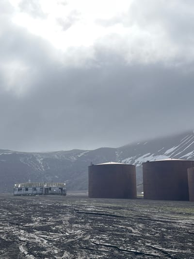 The landscape features large, cylindrical metal structures amidst a harsh environment. Mountains rise behind the structures, partially covered in snow and shrouded in mist. A weathered, rectangular building sits nearby. The ground is dark and textured, suggesting a rugged or desolate terrain. Overcast skies dominate the scene.