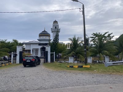 The image shows the exterior of a mosque in Baroh Geunteut, Indonesia, under an overcast sky. A black SUV is parked on a gravel driveway in front of the mosque's entrance. The mosque features a dome and a minaret, with palm trees and other greenery surrounding the structure. A lamppost is visible on the right side of the image. The overall scene is one of a religious building in a tropical or sub-tropical environment.