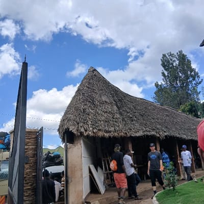 The scene depicts an outdoor gathering in Mweka, Tanzania, under a partly cloudy blue sky. A prominent building with a large, conical thatched roof dominates the right and center of the image. The building features open-sided sections supported by pillars, revealing an interior where several individuals are congregated.

In the midground, a group of approximately five men is visible. One man wears a black shirt, orange shorts, and a dark cap, facing left. Another, in a white t-shirt and dark pants, stands near him. A third man in a dark grey t-shirt and dark shorts stands slightly apart, facing towards the viewer's left. Further under the thatched roof, a man in a blue patterned shirt and another in a white t-shirt with a logo are visible. On the far right, only the shoulder and arm of a person wearing a red garment are visible, suggesting they are in the immediate foreground.

To the left of the building, a tall, dark vertical pillar stands beside a partially open metal gate. Beyond the gate, a white truck or large vehicle is visible, loaded with numerous blue and multicolored bags or luggage. Several people are gathered near this vehicle, partially obscured. White panels or boards are propped against the building's exterior near its entrance. A tall green tree is visible in the background, behind the right side of the thatched roof. Green grass and small plants are present in the immediate foreground to the right.