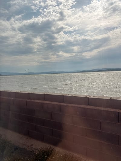 The image shows a body of water with waves under a cloudy sky. In the distance, hills are visible. In the foreground, a brick wall runs horizontally across the bottom of the frame. There are three birds flying in the sky above the water. This is a scenic view of the coastline near Narbonne, France.