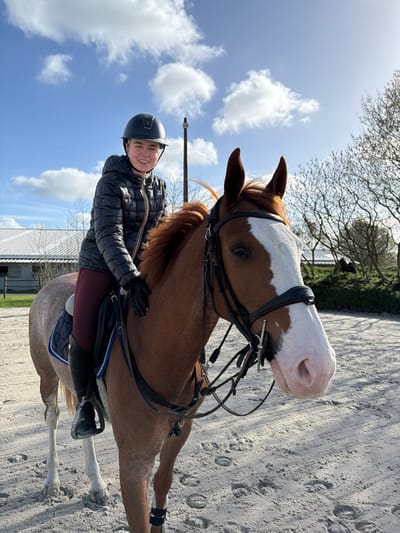 The image captures a young female rider mounted on a chestnut horse within an outdoor equestrian arena in Enghien, Belgium. The rider, facing the camera with a smile, wears a black riding helmet, a black quilted jacket with a small K-Way logo on the chest, dark red breeches, black riding boots, and black gloves. The horse features a prominent white blaze on its face extending to its muzzle, a reddish-brown mane, and lighter lower legs. It is equipped with a dark leather bridle, a snaffle bit, black leg wraps on its front legs, a black saddle, and a dark blue quilted saddle pad. The ground consists of light-colored sand with visible hoof prints. In the background, a large building with a light-colored, multi-peaked roof, consistent with an indoor riding arena or stable complex, is visible to the left. Bare-branched deciduous trees and green shrubbery line the right side of the background. A tall, dark utility pole stands behind the rider. The scene is illuminated by bright sunlight under a blue sky with scattered white cumulus clouds.