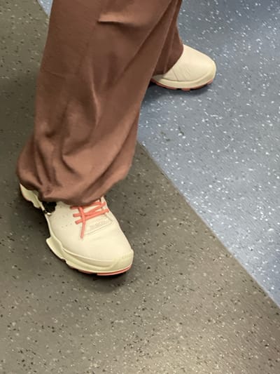 A person is standing on a floor with two distinct patterns. The person is wearing loose-fitting, dusty rose colored pants and white athletic shoes with pink laces. The shoes have a thick, white sole with pink trim around the heel. The floor appears to be a patterned vinyl or linoleum material. The top left portion of the floor is a light blue-gray with small white speckles. The bottom right portion of the floor is a darker gray with speckles of black, white, and what appear to be tiny reflective elements. A diagonal line separates these two floor patterns. The scene is lit by overhead lighting, typical of an indoor environment. There is no visible text in the image. The image appears to be taken from a high angle, looking down at the person's legs and feet.