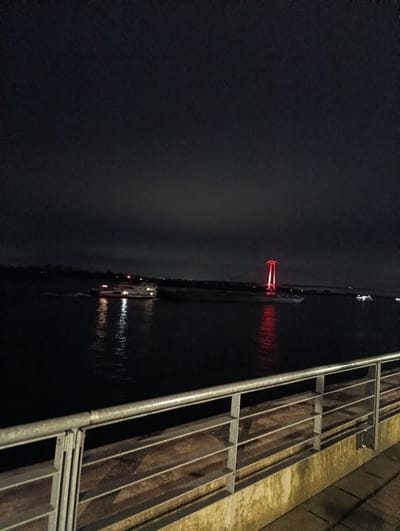 The image depicts a night scene in Emmerich on the Rhine, Germany. A gray metal railing is in the foreground, occupying the lower third of the frame. Beyond the railing is the dark expanse of the Rhine River. On the river, there is a boat with illuminated white lights. Further in the distance, a bridge is visible with red lights at its upper portions, reflecting in the water. The sky is dark, with possible clouds obscuring most of the view. The scene appears to be at night due to the darkness and artificial lights illuminating the subjects.