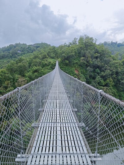 A long, narrow suspension bridge made of metal spans across a verdant valley in Lekhnath, Nepal. The bridge has a grated metal walkway and is bordered by sturdy metal fencing. Dense green forest covers the slopes on either side of the valley, creating a lush, natural backdrop. The sky above is overcast with a layer of clouds. The perspective of the photograph is from the beginning of the bridge, looking down its length towards the distant end.