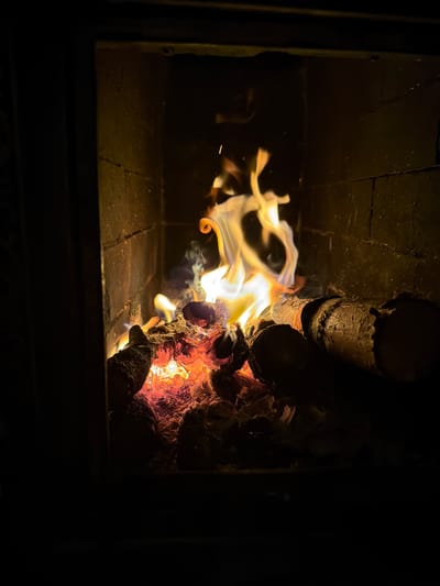 The image shows a fire burning inside a brick-lined fireplace or wood stove. The flames are yellowish-white and swirl around partially burnt logs. The wood on the right side is glowing with orange and red heat. The scene is dark, with the fire providing the main source of illumination. The bricks forming the interior of the stove are visible, showing signs of soot and wear. The location in the city of Пролетарский, Russia is not discernable from the content of the image.