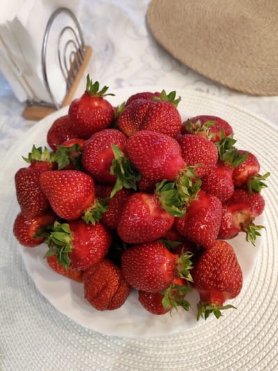 A close-up, overhead shot shows a white plate filled with fresh, ripe strawberries. The strawberries are a vibrant red, with small yellow seeds scattered across their surfaces. Some of the strawberries have green stems and leaves attached. The plate is placed on a textured white placemat. In the background, a metal napkin holder with white napkins is visible, along with a portion of a straw placemat. The scene appears to be indoors, likely on a table. There is no information to suggest this image is located in Kstovo, Russia, or any other specific city.