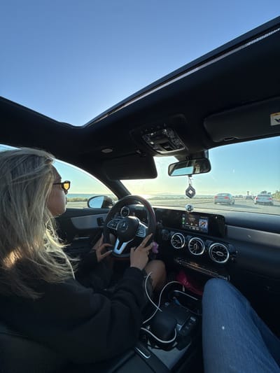 A woman is driving a Mercedes-Benz on a highway in Huntington Beach, California, United States. She is wearing sunglasses and has blonde hair. The car has a sunroof that is open, and several other cars are on the highway. The dashboard and steering wheel are visible. A phone is charging in the center console.