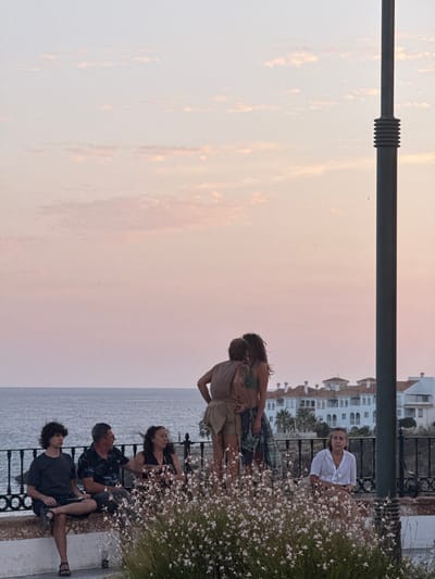 This media file shows a group of people gathered on a balcony overlooking the sea, with buildings in the background. The time of day appears to be late afternoon or early evening, as the sky is a soft pink and orange hue. The weather seems calm and pleasant.

There are several individuals visible in the image. In the foreground, a couple is embracing. Further back, a woman in a white top is seated, looking towards the camera. A group of men are also present, some seated and others standing. One man in a black patterned shirt is looking intently at someone off-camera. Another young man with curly hair is seated and looking down.

The setting is a balcony with a railing, and there is lush greenery with small white flowers growing abundantly. In the distance, white buildings with balconies and roofs are visible, characteristic of Mediterranean architecture. The sea stretches out to the horizon, appearing somewhat overcast.

The overall mood seems relaxed and intimate, with the couple's embrace suggesting affection. The scattered grouping of the other individuals implies a casual social gathering. No visible text is present in the image. The location context of Nerja, Spain, aligns with the architectural style and the seaside setting.