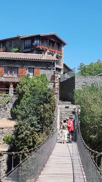The image shows a suspension bridge leading to a stone staircase in the town of Rupit i Pruit, Spain. The bridge is made of wood planks and metal mesh railings. Two people, a woman in a white and yellow skirt and a man in a red t-shirt and black shorts, are standing on the bridge, facing away from the camera and looking up the stairs. The buildings in the background are constructed from stone with tiled roofs and wooden balconies adorned with red flowers. Lush green trees surround the structures, and the sky is clear and blue.