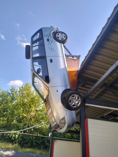 A silver car is suspended upside down from a metal structure, which appears to be part of a car wash facility. The car is positioned at an angle, with its front end pointing upwards and its rear end towards the ground. A portion of a corrugated metal roof is visible above and to the right of the car. The car has a hexagonal pattern design on its side and text that reads "CARWASH TINTILA". The background consists of a bright blue sky with scattered white clouds, and lush green trees. In the foreground, there is a fence with string lights attached, and a darker area that could be asphalt or a paved surface. The scene suggests a playful or artistic display at a car wash in Șoldănești, Moldova.
