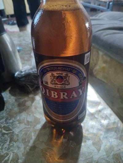 In Bagzar, Uzbekistan, a close-up shot focuses on a bottle of "QIBRAY" branded beverage. The bottle, filled with a golden-brown liquid and topped with a frothy head, rests on a patterned surface. The label prominently displays the brand name "QIBRAY" in large, red letters, framed by a blue and gold emblem. Above this, the text "AJ INTER-ROHAT" and "QK" are visible, along with what appears to be the word "IFTIROR". To the left of the bottle, a person's legs are visible, clad in dark pants, suggesting an indoor setting. The background is blurred, but a grey upholstered surface and hints of other objects are discernible. The lighting suggests an indoor environment, possibly a home or a casual gathering space.