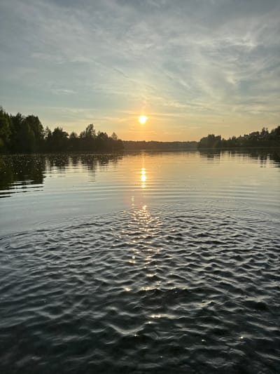This is a landscape photograph taken from the surface of a body of water, likely a lake or river, looking towards the horizon during sunset. The sun, a bright orb with a distinct halo, is positioned slightly left of center in the upper portion of the image, casting a warm golden light across the scene. The sky is a gradient of soft blues and yellows, with wispy clouds that are illuminated by the setting sun.
In the middle ground, a line of trees forms a dark silhouette against the bright sky, marking the distant shore. The water in the foreground is choppy, with small waves creating ripples and reflecting the sunlight. There are no visible people, structures, or animals in the image. The overall impression is one of tranquility and natural beauty.
The city of Kamysh, Russia, is known for its numerous lakes, particularly the Kamyshla lake, which is a popular recreational area. Given the natural landscape and the presence of a lake, this image could have been taken at or near Kamyshla lake.