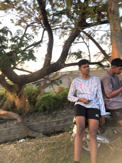Two young males are seated on a motorbike, with one standing in front of the other. The male in the foreground is wearing a light blue long-sleeved shirt with a white and red lightning bolt pattern, black shorts, and holding a black smartphone in his right hand. The male behind him is wearing a grey t-shirt. Both males appear to be of Asian descent. The motorbike is black and silver. They are positioned outdoors near a large tree with thick branches. The background consists of foliage and what appears to be a concrete channel. The lighting suggests it is late afternoon or early evening, with sunlight filtering through the trees, casting long shadows and creating a warm glow. The scene is slightly out of focus due to motion blur. The location is identified as Sentul, Indonesia, suggesting a possibly rural or suburban environment on the outskirts of a city.