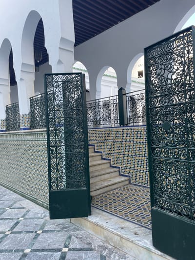 The scene depicts an architectural space within the city of Moulay Idriss Zerhoun, Morocco, likely an entrance or courtyard of a traditional building. In the immediate foreground, two dark green, ornate wrought-iron gates, featuring intricate scrollwork patterns, stand ajar, extending towards the viewer. The ground surface in front of the gates is composed of large, light-grey marble tiles interspersed with darker square inserts, forming a checkered pattern.
Beyond the gates, a short flight of five light-colored marble or stone steps ascends towards the right. The walls adjacent to these steps and forming a raised platform are covered in traditional Moroccan zellige mosaic tiles, patterned with small blue, yellow, and white squares, bordered by a solid blue line at the base. Along the edge of this elevated area, a dark green, wrought-iron railing with scrollwork identical to the gates is present, supported by dark, square pillars.
In the background, a white architectural structure with a series of five distinct, white horseshoe arches supported by thick white pillars forms an arcade or colonnade. Beneath these arches, a dark wooden ceiling with visible beams is apparent. A single, dark, bell-shaped lantern hangs from the ceiling within one of the archways on the left. Through and beyond the arches, additional white buildings are visible, some featuring subtle green architectural details, possibly indicating roof tiles. No people or animals are present. The scene is illuminated by natural daylight.