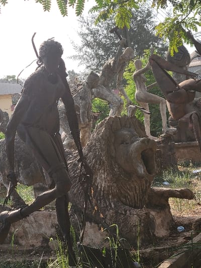 This image displays an outdoor sculpture collection located in San Martín, Colombia, under bright daylight. The foreground is dominated by a dark, metallic-toned sculpture of a human figure in a striding pose, possibly constructed from assembled metal, holding an elongated object that resembles a spear. To its right, a large, light-colored sculpture of a roaring lion's head and upper torso emerges from a rocky base, its mouth agape. Behind the lion and the foreground figure, several other light-colored sculptures are visible: one human-like figure is depicted mid-swing with a hammer-like tool, another is a crouching nude female figure, and a larger, abstract, possibly insectoid sculpture is partially visible on the far right.
The sculptures are situated on uneven, earthy ground sparsely covered with green grass and weeds. Some discarded items, including a clear plastic bottle, are present on the ground. The background features various green trees and shrubs. On the far left, a light-colored building with a corrugated metal roof is partially visible. Overhead, green leafy branches from trees partially frame the top of the scene against a bright, overcast sky.