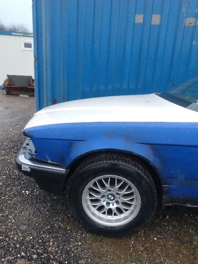 The image displays the front left portion of a two-tone vehicle, situated on a wet gravel surface in Bitburg, Germany. The vehicle features a white hood and a blue front left fender, which shows a visible dent or crease above the wheel well and is splattered with mud or grime. A chrome bumper with black trim and a possible sticker is present at the front lower section. The front left wheel is equipped with a silver multi-spoke alloy rim, centered with a BMW logo, and fitted with a black, wet tire exhibiting a pronounced tread pattern. In the background, a large, corrugated blue metal structure, likely a shipping container, stands vertically to the right of the vehicle. To the left of the blue structure, a white modular building with a rectangular window and a partially visible "Condecta" sign is present. Further in the background, bare trees are visible under an overcast sky. The ground consists of dark, wet gravel with some patches of green vegetation. No individuals are present in the scene.