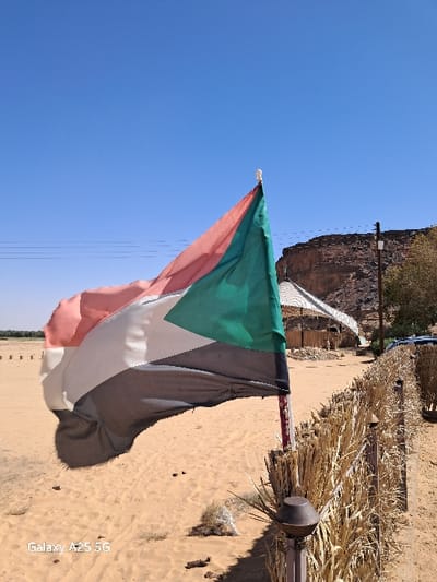 The image shows the flag of Sudan flying in the wind in front of a tall, brown, rocky hill. The flag is attached to a pole and is secured to a fence made of woven, dry grass. The ground is a sandy, light brown color. The flag is waving in the wind and there is a clear blue sky in the background. The photo was taken near the coordinates 18.54 latitude, 31.831 longitude, which is located in Karima, Sudan. The photo was taken during the daytime, as the sun is shining and the sky is clear. There is a small, white building on the right side of the photo. The image was taken with a Samsung Galaxy A25 5G phone. There are no people in the image.