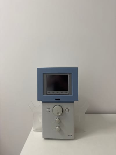 The image shows a medical device, likely a physiotherapy or rehabilitation unit, mounted on a white wall. The device has a blue frame around a screen displaying what appears to be a grid or graphical interface. To the right of the screen, there are several control knobs and buttons. Some text is visible on the device, including "ESC," "enter," "press to start/stop," "time," and "intensity." There is also a logo with three interconnected squares, possibly representing a brand or manufacturer. The device is supported by clear plastic brackets attached to the wall. The background is a plain white wall, with a hint of a white ceiling or upper wall at the top of the frame. The lighting suggests an indoor setting, possibly a clinic or medical office. There are no people visible in the image, and no explicit indication of the time of day or weather. The overall impression is that of a functional piece of medical equipment in a sterile or clinical environment, located in Suceava, Romania.
