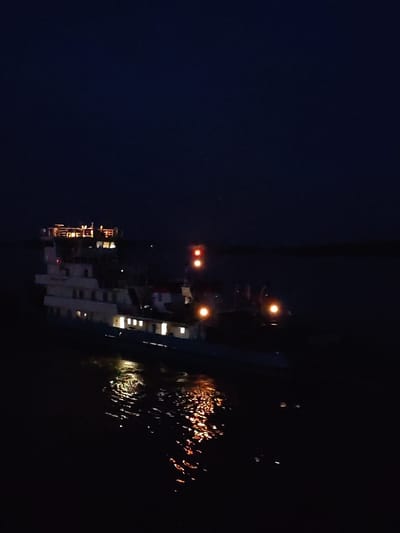 A multi-decked vessel is illuminated by its internal lights and external running lights as it navigates through dark water at night. The vessel is a light color, possibly white or cream, with several decks visible, each featuring rows of illuminated windows. The top deck appears to have an open area with railings and additional lighting. Red running lights are visible on the vessel's structure. The water reflects the ship's lights, creating shimmering patterns. The sky is dark and cloudless. The scene is captured from a slight elevation, looking down at the vessel. The location is identified as Kudryashovo, Russia.