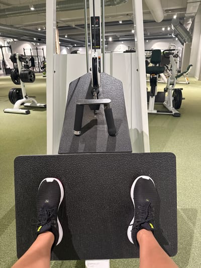 The image captures a first-person perspective of a gym interior, focusing on the user's feet and a piece of exercise equipment. The scene is brightly lit, likely indoors, with a green carpeted floor and white walls or partitions. Overhead, silver ventilation ducts and numerous lights are visible, indicating a modern facility.
In the foreground, a person's feet, clad in black and white Puma athletic shoes with yellow accents, rest on a textured black platform. This platform is part of a larger gym machine, predominantly white with black handles and components. The visible machine setup suggests it could be a seated row or a similar strength training apparatus where the user pushes or pulls.
In the background, a variety of other gym equipment is discernible, including weight stacks, benches, and various machines, some with green upholstered pads. Weight plates are neatly stacked on a rack further back. The overall setting is that of an active and well-equipped fitness center.
Visible text includes "PUMA" on the user's shoes and "PRECOR" and "IMC" on a label attached to the white gym machine in the mid-ground. The image portrays an individual engaged in or preparing for a workout session.
