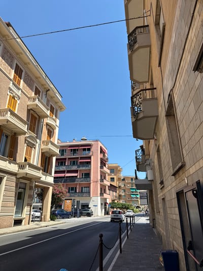 The image captures a street scene in Finale Ligure, Italy, on a clear, sunny day. The perspective is from a street level, looking down a road flanked by multi-story buildings. On the left, a building with ornate detailing and orange shutters is visible. The right side of the street is dominated by a building with a facade of light-colored bricks and balconies with decorative black railings. Several cars are parked along the road, including a blue SUV and a white hatchback. Pedestrians are not clearly visible, but a person on a bicycle is seen further down the street. The sky is a vibrant blue. A utility wire stretches across the upper portion of the frame.