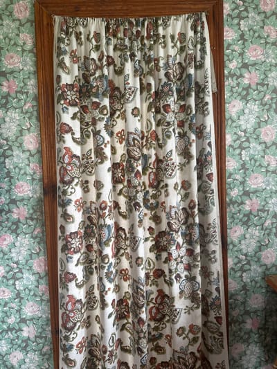 A floral-patterned curtain is positioned inside a wooden frame, set against a background of floral wallpaper. The wallpaper features pink and white flowers against a green backdrop. The curtain has a dense floral print on a white base, with shades of brown, red, green, and blue. The curtain is visibly pleated and slightly bunched within the frame.
The image does not contain the necessary information to determine the location in the city of Студзёнкаўскі сельскі Савет, Belarus.