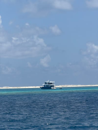 The scene depicts a maritime landscape in Gulhi, Maldives. The sky is a light blue with scattered white cumulus clouds. The foreground consists of deep blue, choppy ocean water with subtle sun reflections. In the midground, a multi-decked liveaboard vessel, featuring a dark blue hull and white superstructure, is stationary in a stretch of vibrant turquoise water. To the immediate right of this vessel, a smaller, dark-colored dinghy is also present in the turquoise water. Beyond the vessels and turquoise water, a narrow, elongated white sandbank or shallow reef crest extends horizontally across the frame. No individuals are visible on either vessel or the sandbank.