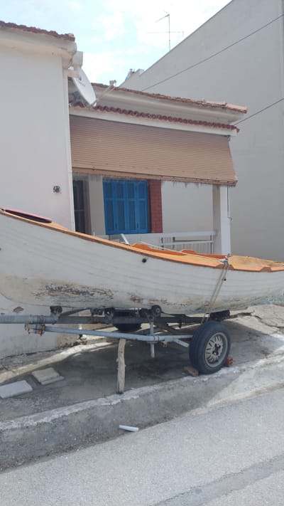 A white rowboat with an orange trim is parked on a trailer outside a building with a white facade and a brown retractable awning over blue shutters. The boat appears weathered, with peeling paint on its lower hull. The trailer has a single tire and is supported by a wooden post. In the background, a satellite dish is visible on the roof of an adjacent building, along with an antenna. The scene is located in Νέα Μηχανιώνα, Greece, on a paved surface next to a road. The sky is partly cloudy.