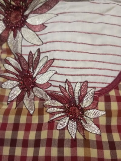 The image shows a garment featuring three embroidered, stylized flowers with beaded centers. The petals are two-toned, with one side a deep red and the other a light silver, outlined in black thread. The background consists of two distinct fabrics: one is a white fabric with thin red horizontal stripes, and the other is a red and tan checkered pattern. A red binding trims the top edge of the garment where the fabrics meet.