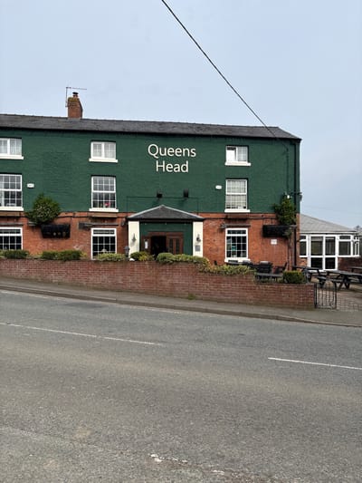 An exterior forensic view captures "The Queens Head" public house in West Felton, United Kingdom, under an overcast sky. The two-story building features a dark green textured facade on the upper level and red brickwork on the lower level. White text spelling "Queens Head" is centered on the green facade. Numerous white-framed windows are distributed across both levels. A dark grey tiled roof, a brick chimney, and a television antenna are visible at the top. The central main entrance has a small dark canopy and double wooden doors, flanked by two wall-mounted lights. A low red brick wall with green shrubs runs along the building's frontage. Outdoor seating, consisting of dark tables and chairs, is situated behind this wall. To the right, a single-story white-framed conservatory-style extension with additional picnic tables is visible. The foreground consists of an asphalt road sloping slightly upwards, marked with a faded white dashed center line, separated from the building by a narrow grey curb. No individuals are present. A single dark utility line traverses the upper right portion of the frame.