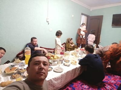 The image depicts an indoor family gathering within a residential setting in Samarkand, Uzbekistan. The room features light blue textured walls, white crown molding, and a dark wooden door, slightly ajar, revealing a light-colored curtain. A flat-screen television is mounted on the far right wall. The floor is covered with a vibrant red, blue, and black patterned rug, consistent with Central Asian interior design.
A low table, draped in a white tablecloth, serves as the central point for a communal meal. It is laden with a diverse spread including a prominent large plastic bottle of orange soda, several pieces of traditional flatbread (non), plates of nuts, dried fruits, various snacks, and fresh fruit such as green grapes, red apples, and a lighter-colored melon or pomelo. Numerous small plates, bowls, and drinking glasses are distributed across the table.
In the immediate foreground, a man in a dark grey long-sleeved shirt is taking a selfie, looking directly at the camera. To his left, the face of another man is partially visible. Further back on the left, a man in a dark blue long-sleeved shirt is seated on a low cushion or mat against the wall.
On the right side of the table, a child in a dark blue hooded sweatshirt sits with their back to the camera. In the background, a young child in light pink pajamas stands near the open door. A woman in a patterned brown and orange dress, holding an infant wrapped in a light-colored blanket, stands near the door. To her left, an older woman, possibly wearing a headscarf, is seated on the floor in patterned yellow-brown clothing. A partially visible individual wearing an orange and black patterned garment is on the far right, hunched over. The scene indicates a multi-generational social gathering or family meal.
