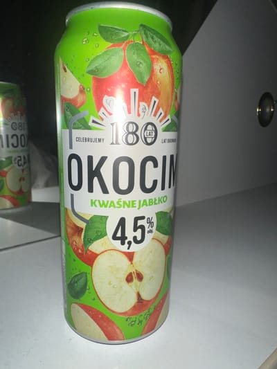 The image shows a can of Okocim Kwasne Jablko, a Polish brand of apple-flavored beverage. The can is predominantly green with illustrations of apples, leaves, and water droplets. The brand name "OKOCIM" is prominently displayed in large black letters. Below it, "KWAŚNE JABŁKO" (sour apple) indicates the flavor. To the left, there is a circular logo with the number "180" and text that reads "LAT BROWARU" (years of brewery), suggesting the brand's long history. Further text on the can includes "CELEBRUJEMY" (we celebrate) and "4,5% obj." (4.5% alcohol by volume). The can is positioned horizontally and slightly angled, with a reflection visible below, suggesting it is placed on a reflective surface. The background appears to be a neutral indoor setting, possibly a shelf or countertop, with a hint of overhead lighting. There are no people or a specific event depicted; the focus is solely on the product packaging.