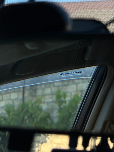 The image captures an interior view from a vehicle, likely a car, looking out through the side window. The scene is dominated by a WeatherTech sticker affixed to the window, oriented vertically. The sticker is blue and white with the text "WeatherTech" prominently displayed in a stylized font. Below this, smaller text is visible, possibly a website or phone number, though it is too blurry to read clearly.
Outside the window, a blurred backdrop suggests an outdoor environment. Green foliage and what appears to be a textured surface, possibly a stone wall or building, are discernible. The lighting suggests it is daytime, with sunlight casting reflections on the window and the car's interior.
The foreground is dark and out of focus, showing the car's interior structure, including a textured fabric on the left and a dark, smooth surface on the right, possibly part of the dashboard or door panel. The overall impression is a snapshot taken from within a moving or stationary vehicle, focusing on the window and its accessory. The location context of Mastaga, Azerbaijan, does not provide specific visual cues within the image itself.