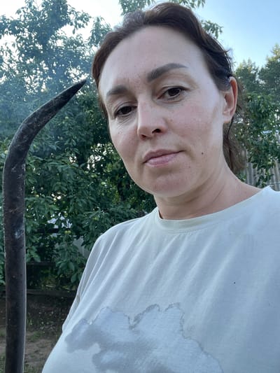 A close-up, selfie-style photograph shows a woman in her late 30s or early 40s from the chest up, against a backdrop of greenery and a rustic fence. The woman has medium brown hair pulled back and is looking directly at the camera with a neutral expression. Her skin appears slightly flushed and there are visible pores and fine lines. She is wearing a light-colored t-shirt, which has dark, damp patches on the chest area, suggesting she may have been sweating. To the left of the woman, a portion of a curved, dark metal object, possibly a scythe or sickle, is visible. The background consists of lush green foliage, likely from trees and bushes, with some small green fruits appearing on the branches. Behind the greenery, a weathered wooden fence and a glimpse of a gray, corrugated metal structure can be seen. The lighting suggests it is daytime, possibly late afternoon, with a soft, natural light illuminating the scene. The overall setting appears to be an outdoor, possibly rural or garden environment, in Bormist, Russia.