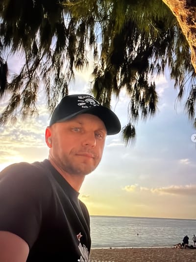 A man wearing a black t-shirt and a black baseball cap is taking a selfie outdoors. He has blue eyes and short, light brown hair. He is wearing a white earbud in his left ear. The baseball cap has a design on it. The man is looking directly at the camera. Behind him, there is a body of water, likely the ocean, with a sandy beach in the foreground. The sky is clear with some clouds, and the sun appears to be setting or rising, casting a warm glow over the scene. There are some trees with long, wispy leaves hanging from the top of the frame, creating a natural canopy. In the distance, on the beach, a few other people can be seen. The man's left arm is visible in the foreground, resting on his chest. The scene appears to be a serene beach environment, possibly during golden hour. This image is not located in the city of Ban Phonsavang Neua, Laos.