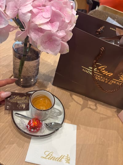 The image shows a table setting with various items related to Lindt chocolate, a luxury Swiss chocolatier with its main factory and headquarters in Kilchberg (ZH), Switzerland. A dark brown Lindt shopping bag is visible in the background, and a Lindt napkin rests on the right side of the table. A small transparent glass cup of coffee sits on a saucer, along with a silver spoon and a red Lindor chocolate truffle. A brown Lindt chocolate Café packet is placed near the cup. In the lower left corner, pink hydrangeas in a silver patterned vase add a touch of elegance. A hand with a manicured nail is visible near the coffee. The table appears to be light-colored wood.