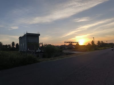 The image depicts a road scene in Cotbuket, Indonesia, under a sunset. A two-story concrete building stands to the left, partially obscured by vegetation. In the background, there is a simple wooden structure, possibly a shelter or carport, along with several houses and trees. A motorcycle is visible on the right side of the road. The sky is filled with streaky clouds, and the sun is low on the horizon, casting a warm glow. The road surface is dark and appears to be asphalt.