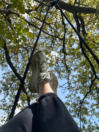 The image shows a person's foot in a sneaker resting on a thick, dark rope, possibly part of a rope bridge or climbing structure. The leg is clad in dark gray or black sweatpants. Above, a tree canopy with green and yellowing leaves is visible against a pale blue sky. The location is Liava Reka, Bulgaria.