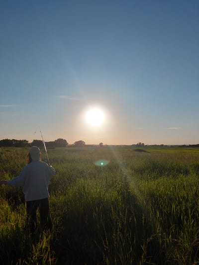 A person wearing a light-colored baseball cap and a light-colored long-sleeved shirt stands in a field of tall green grass, holding a fishing rod. The person is facing away from the camera and towards the horizon. The sun is high in the sky, casting a bright light and creating a lens flare. The sky is a clear blue with some subtle gradients. In the distance, there are trees and a flat landscape that appears to be a field or plains. The image is taken outdoors, likely during the daytime or late afternoon. The exact location in Vozdvizhenka, Russia cannot be determined from the visual information provided.