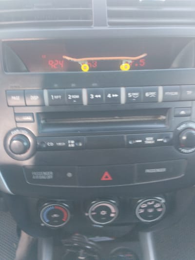 A close-up, slightly blurry, eye-level indoor shot captures the dashboard of a car, likely in Zavolzhye, Russia, as indicated by its Russian context. The primary focus is the car's audio system and climate controls. Dominating the upper portion of the frame is a digital display showing the time "9:24" and what appears to be "7.5" on the right, possibly indicating a radio frequency or another setting. Perched atop this display is a small, toy skateboard with orange decks and yellow wheels. Below the display, buttons and knobs for the car's stereo system are visible, including "PAGE," "DISP," "1 RPT," "2 ROM," "3 4->," "5 DISC," "6 DISC," and "TUNE/SCAN." A central slot for a CD is also present. To the left, a volume knob labeled "PWR/VOL" is prominent, and to the right, a smaller knob is visible. Further down, climate control dials with red and blue indicators are situated, along with symbols indicating airflow direction. A red hazard light triangle is visible in the center console. Text indicating "PASSENGER AIR BAG OFF" is present on the dashboard. The overall impression is a utilitarian car interior, with the toy skateboard adding an unexpected, personal touch.