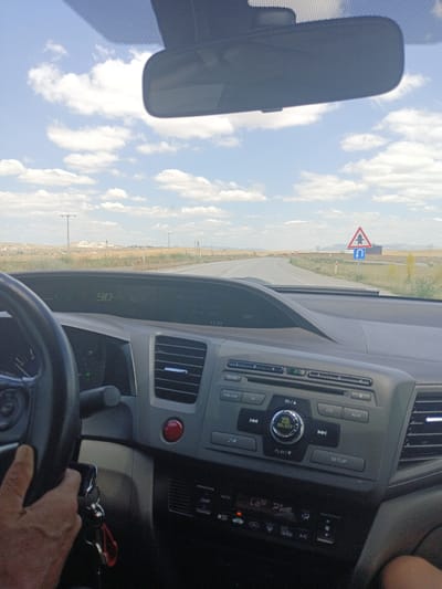 The image shows the interior of a car driving on a road near Balâ, Turkey. The driver's hand is on the steering wheel, and the dashboard is visible, displaying a speed of 90. The road ahead curves to the left, and there are fields and hills on either side. A road sign indicating a U-turn is visible on the right. The sky is blue with some clouds.