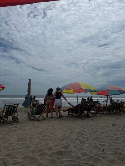 The image shows a beach scene in Montañita, Ecuador, taken at coordinates -1.825 latitude, -80.754 longitude. The beach is mostly empty with a few people present. The scene is dominated by a blue sky with scattered white clouds. The water is calm, with gentle waves lapping on the shore. Two women are walking on the sandy beach, holding hands. They are both wearing swimwear, and one is carrying a red and yellow sarong. In the background, there are several beach umbrellas with colorful stripes, set up on the beach. Several people are sitting in chairs under the umbrellas, with a few others standing around. Some people are playing beach volleyball, and others are sitting on the sand. The scene is one of peaceful relaxation.