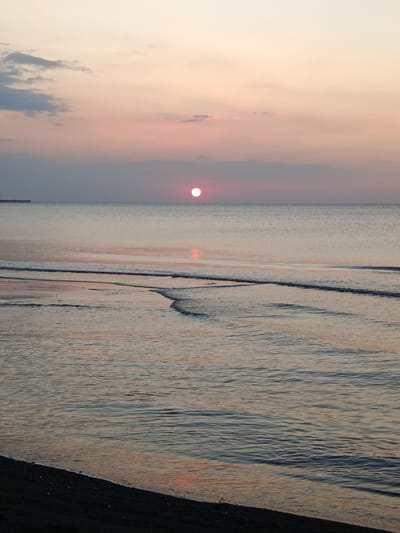 The image depicts a serene seascape at dusk, likely near Jangkar, Indonesia, given the provided context. The sun, a bright pink orb, is low on the horizon, casting a soft glow across the tranquil water. The sky is a gradient of pale orange and pink, with a few wispy clouds. A distant pier or jetty is visible on the left side of the frame, extending into the sea. The foreground shows gentle waves lapping onto a dark, pebbly shore. The overall impression is one of peacefulness and the beauty of nature.