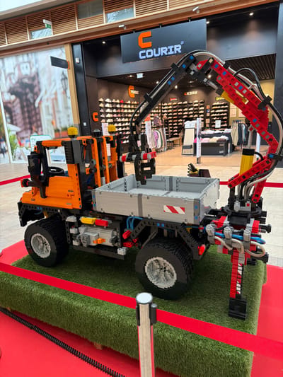 The image captures a detailed, large-scale LEGO Technic model of an orange and grey utility truck with an articulated crane, situated indoors within a commercial space in Mont-Saint-Martin, France. The model features an orange cab with yellow warning lights, a light grey flatbed equipped with red and white diagonal hazard markings, and a prominent red and black crane arm extending from the rear, terminating in a gripper mechanism. The truck model is displayed on a raised rectangular platform covered in artificial green grass, cordoned off by a red rope barrier secured by silver-topped stanchions. In the background, the entrance to a "COURIR" retail store is visible, marked by its orange 'C' logo on a black facade. The store interior showcases numerous rows of athletic shoes on display shelves and racks of clothing. To the left of the store entrance, a large poster on the wall depicts an urban street scene. No individuals are present or interacting with the scene.