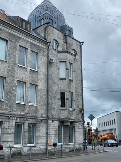 The image depicts a street scene in Sortavala, Russia, under an overcast sky. A multi-story building with a textured grey facade and a distinctive grey domed roof dominates the left side of the frame. The building features numerous windows, some rectangular and others arched. Below the main structure, the lower section is clad in light grey brickwork. A metal fence runs along the sidewalk in front of the building.
On the right side of the image, a white building with a row of rectangular windows is visible. Between the two buildings, an orange tanker truck and a dark sedan are on the road. A person wearing a backpack walks along the sidewalk towards the road. Various electrical wires crisscross the sky. The overall impression is one of an urban environment with mixed architectural styles.