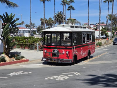 A red and white trolley, labeled "San Clemente Trolley," travels down a street in San Clemente, California. The trolley is a vintage-style vehicle with large windows and a white roof. It is red with a black stripe running along the side. The trolley has the "SF" logo on the front and "San Clemente Trolley" written on the side. The trolley is traveling on a paved road with a bike lane marking. Palm trees line the street, and there are buildings and houses in the background. The sky is blue and clear. The trolley is facing right, and the front of the trolley is visible. The trolley is in motion, and its wheels are visible. The trolley appears to be carrying passengers, as some people are visible through the windows. The trolley is in good condition and appears to be well-maintained. The overall scene is one of a sunny day in a coastal city.