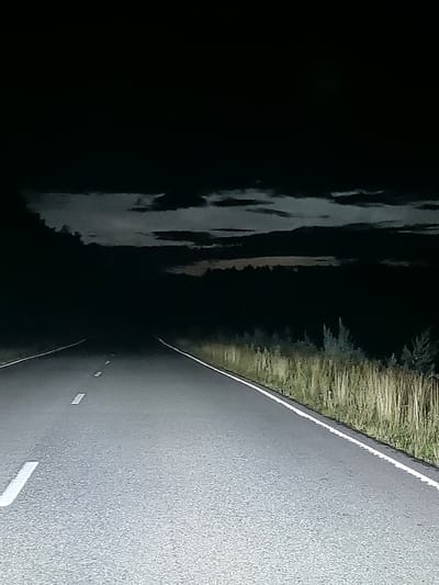 The image shows a desolate road at night, possibly in the vicinity of Velikiy Dvor, Russia. The road is paved and has white lines indicating lanes and dashed lines in the center. The sky above is dark with some visible clouds. The surrounding environment is dimly lit, with tall grass and some sparse trees visible along the side of the road. The darkness and isolation suggest a rural or remote setting. There are no people or other visible subjects in the image, focusing solely on the road and its immediate surroundings.